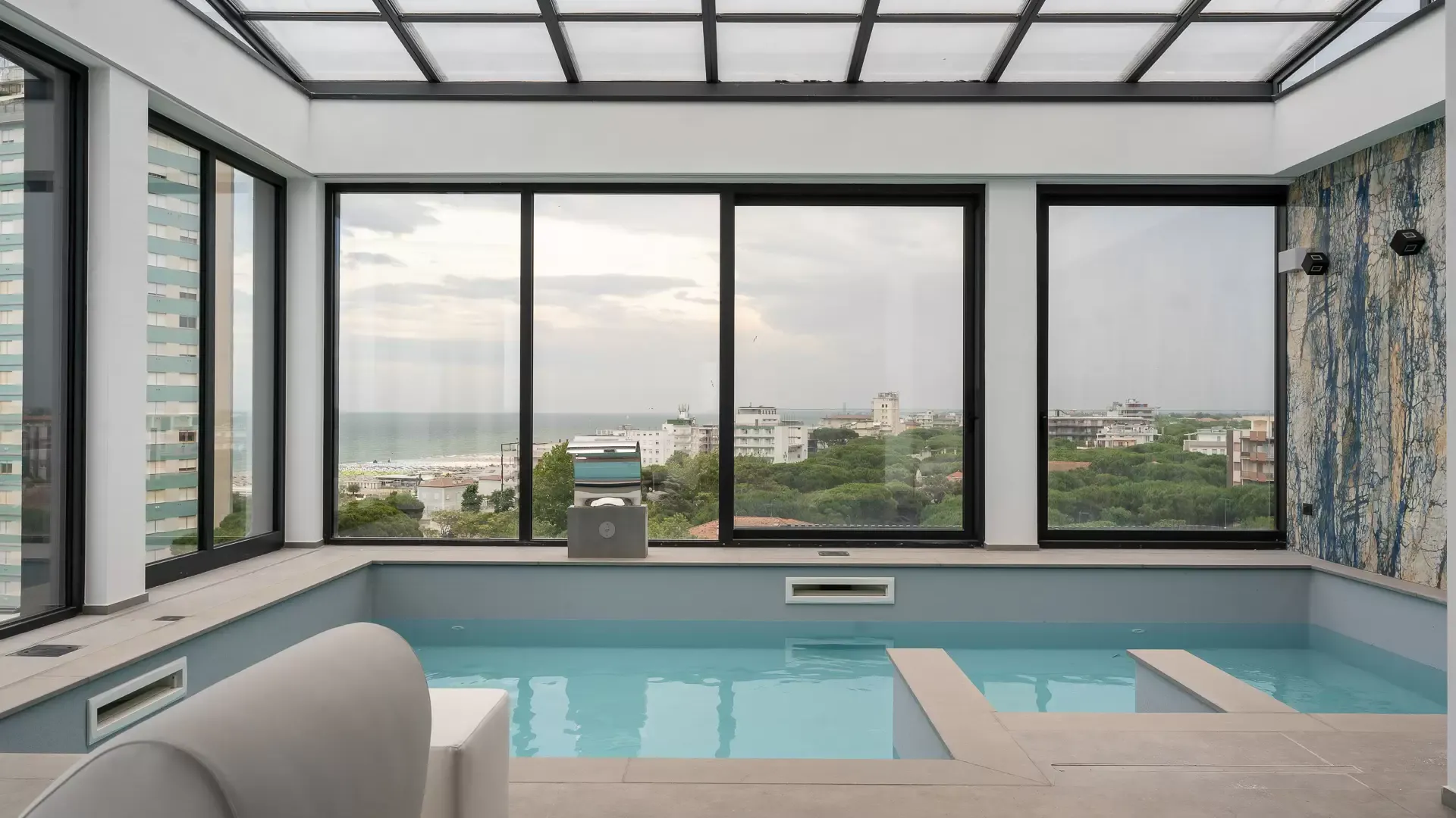 Piscina interna con vista panoramica su città e mare, attraverso ampie finestre.