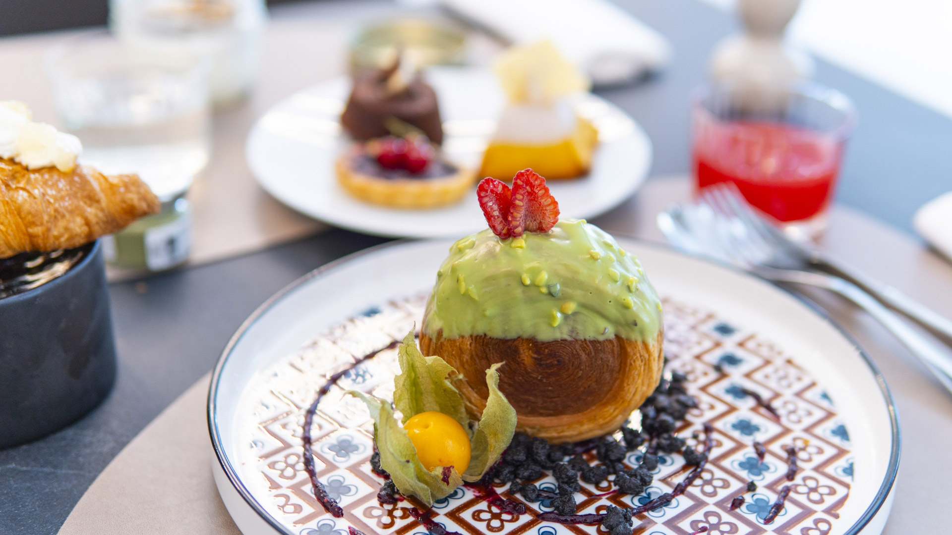Dolci raffinati su piatti decorati: croissant, tortine e un dolce con glassa verde e frutti.
