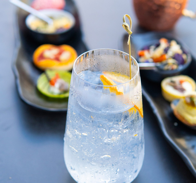 Bicchiere di cocktail rinfrescante con arancia, piatti colorati sullo sfondo, atmosfera elegante.