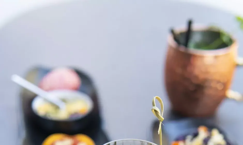 Cocktail con ghiaccio e fetta d'arancia, con stuzzichini sullo sfondo.