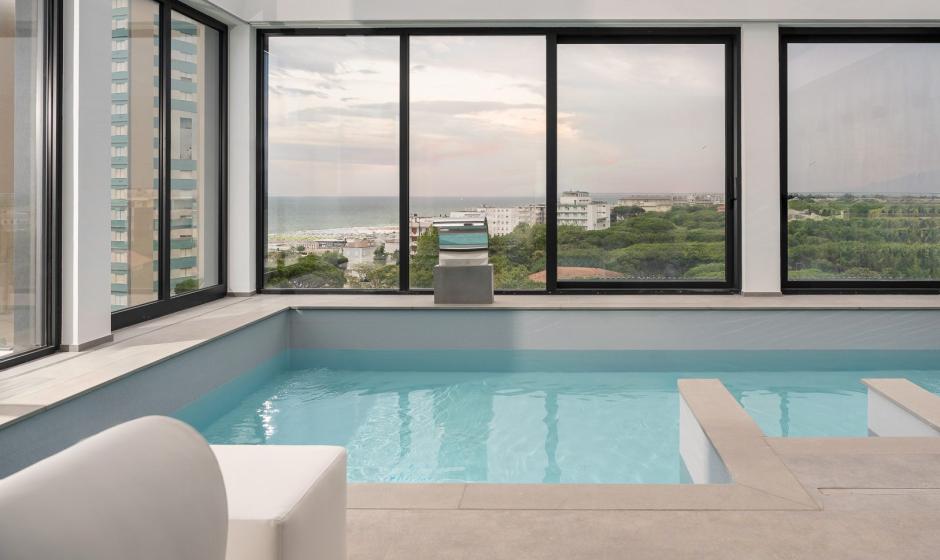 Piscina interna con vista panoramica su città e mare attraverso ampie finestre.