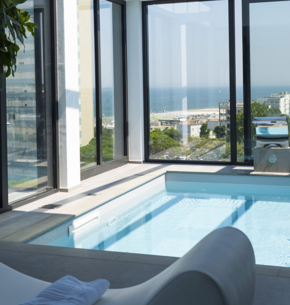 Piscina interna luminosa con vista mozzafiato sul mare e città, atmosfera rilassante e lussuosa.