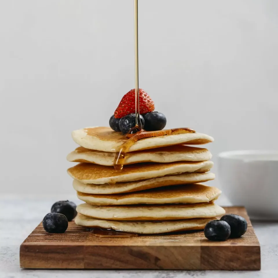 Pila di pancake con frutti di bosco e sciroppo su un tagliere di legno.